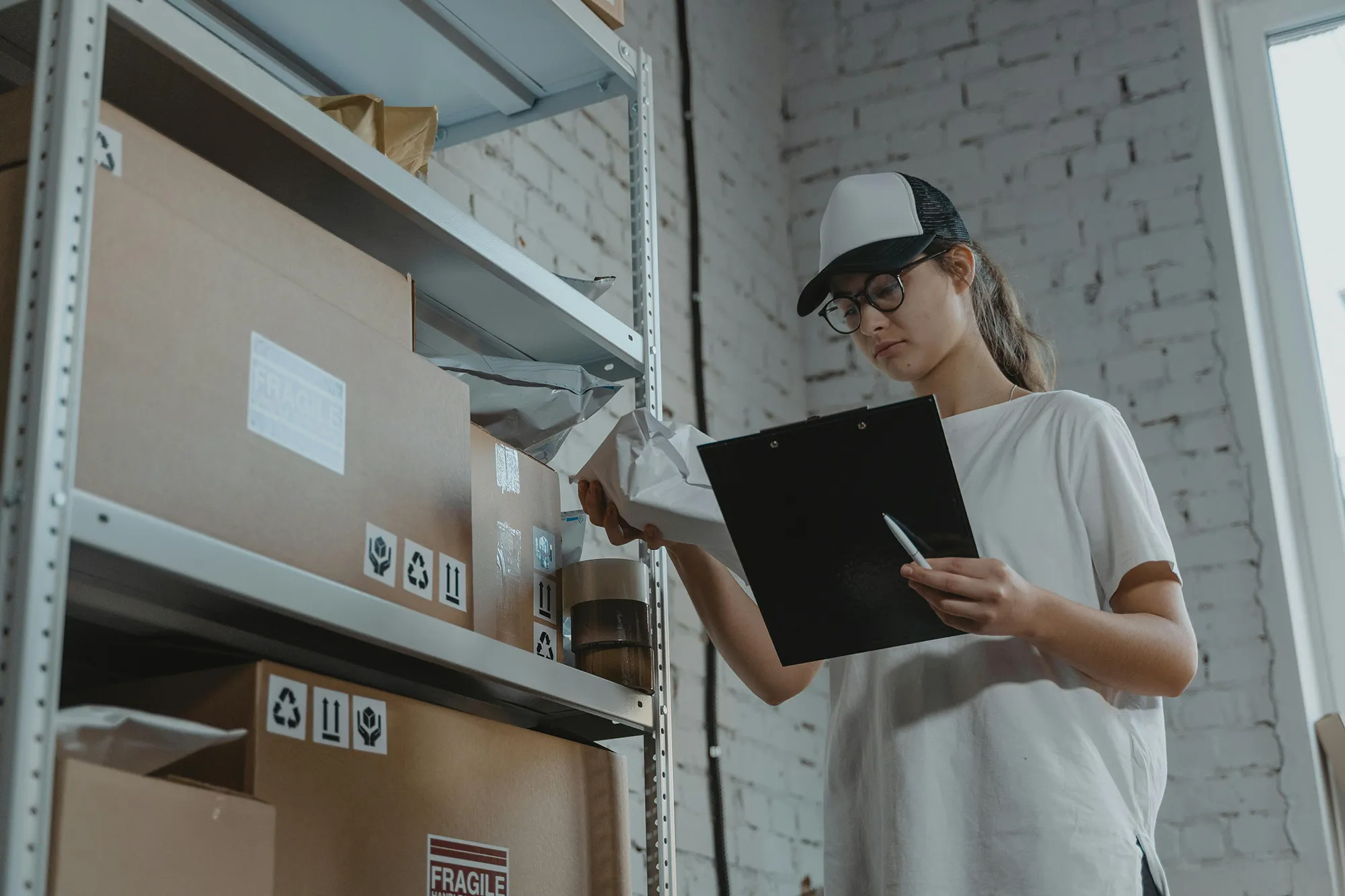 Junge Frau mit Brille und Basecap überprüft Lagerbestände mit einem Klemmbrett vor Regalen voller Kartons.