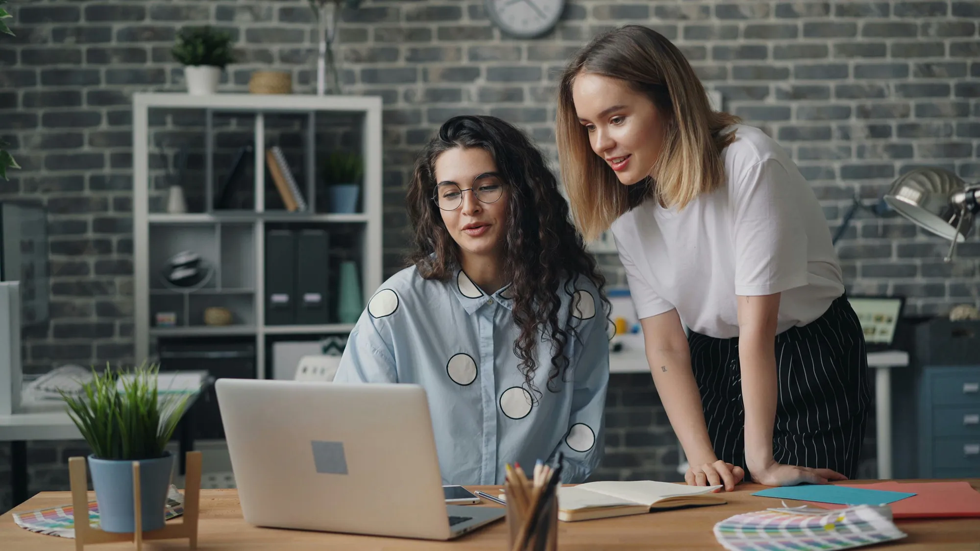 Zwei Frauen in einem modernen Büro arbeiten gemeinsam an einem Laptop, umgeben von kreativen Arbeitsmaterialien.