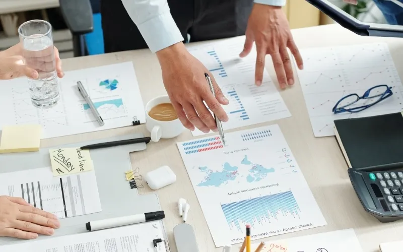 Teammeeting mit Business-Analysen: Hände zeigen auf Finanzdiagramme und Statistiken auf einem Bürotisch mit Laptop, Notizen und Kaffee.